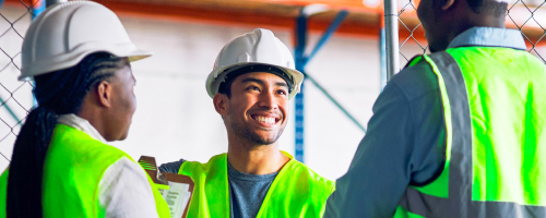 Tres trabajadores con chalecos de seguridad y cascos sonriendo en un almacén